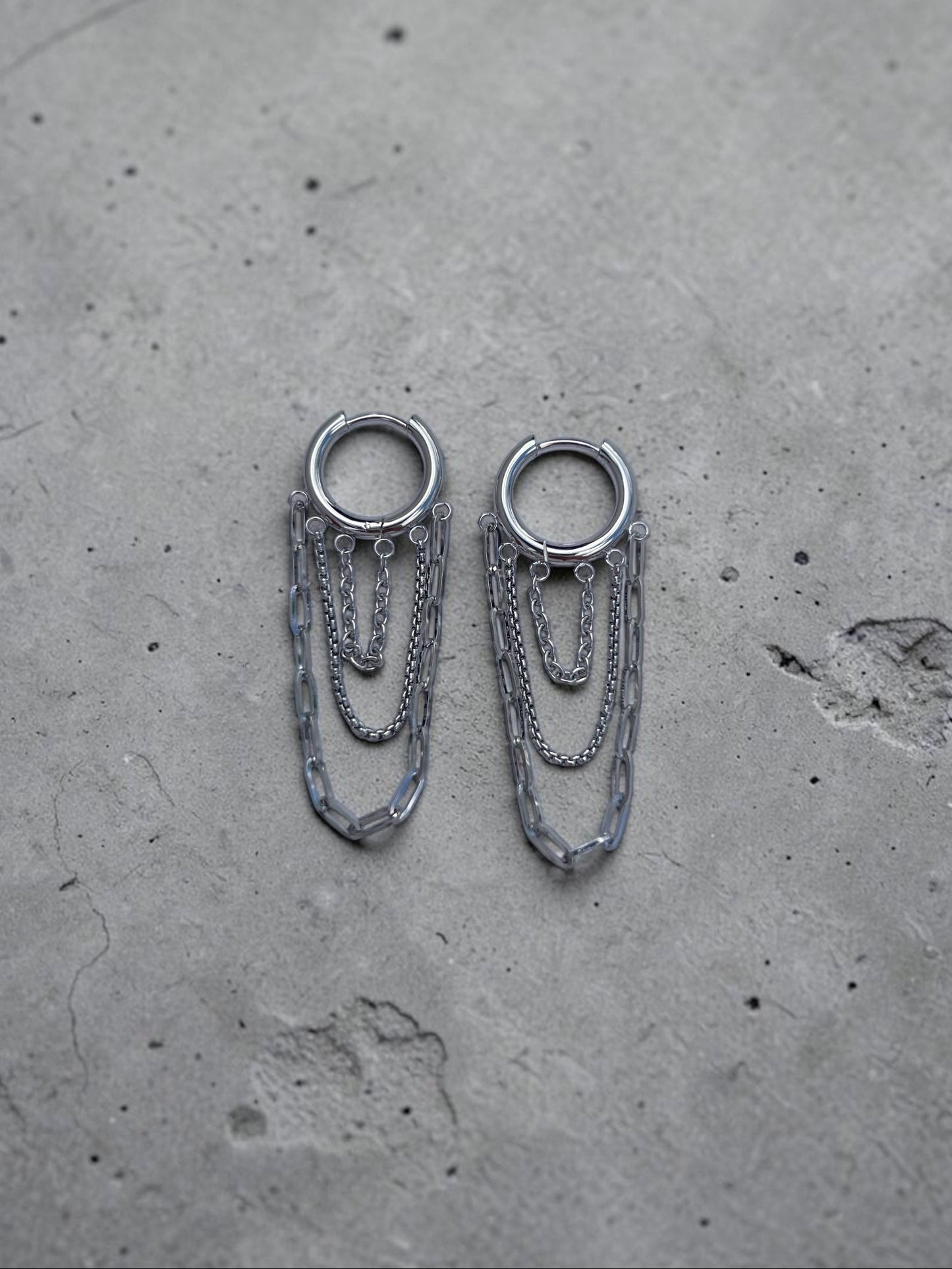 Pair of silver hoop earrings with chain extensions on a gray concrete surface